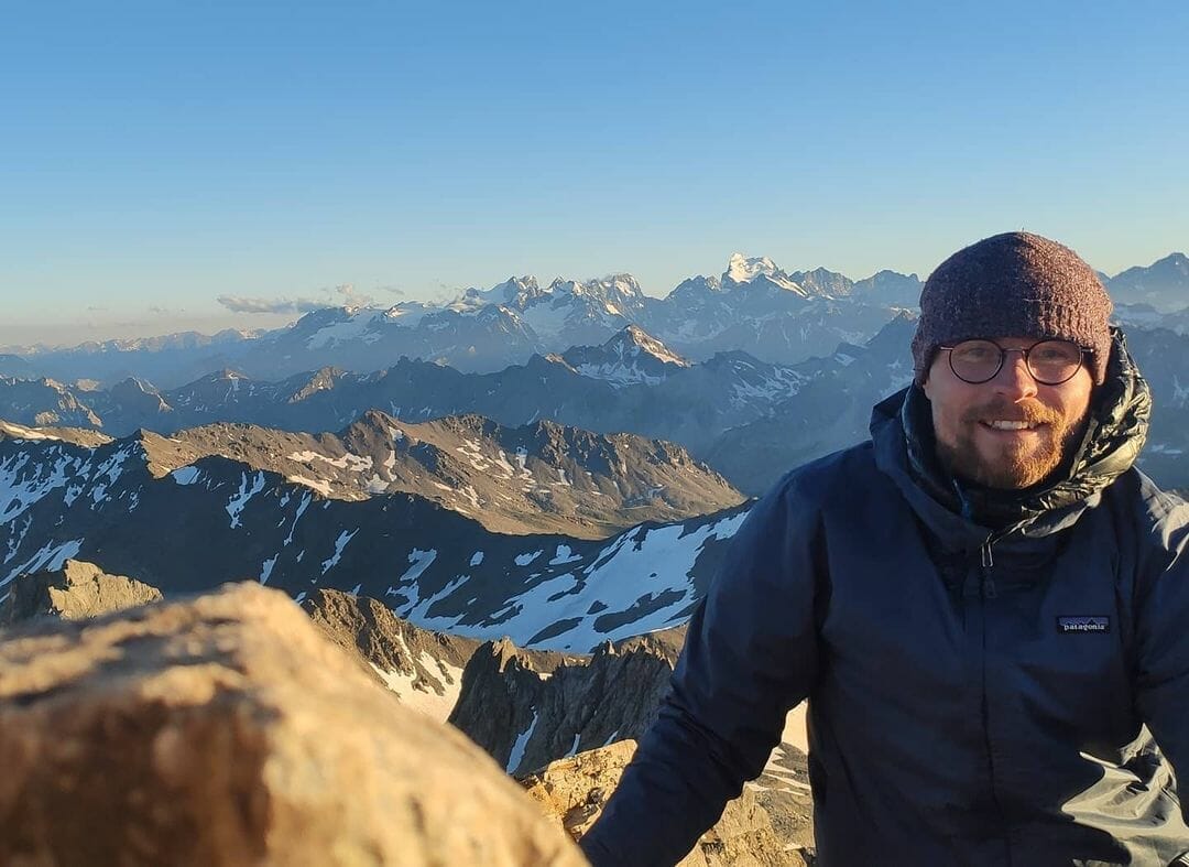 Un homme qui porte un bonnet sourit à la caméra depuis le sommet d'une montagne des Alpes